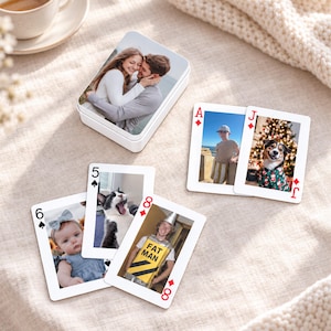 May include: A white rectangular tin box featuring a photo of a couple embracing, accompanied by five playing cards. The cards display various personal photographs, including a baby, a cat, a person in a costume labeled FAT MAN, a child, and a dog.
