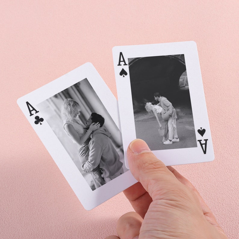 May include: Two black and white photographs of couples printed on playing cards. The cards are the Ace of Spades and the Ace of Clubs. The cards are held in a hand.