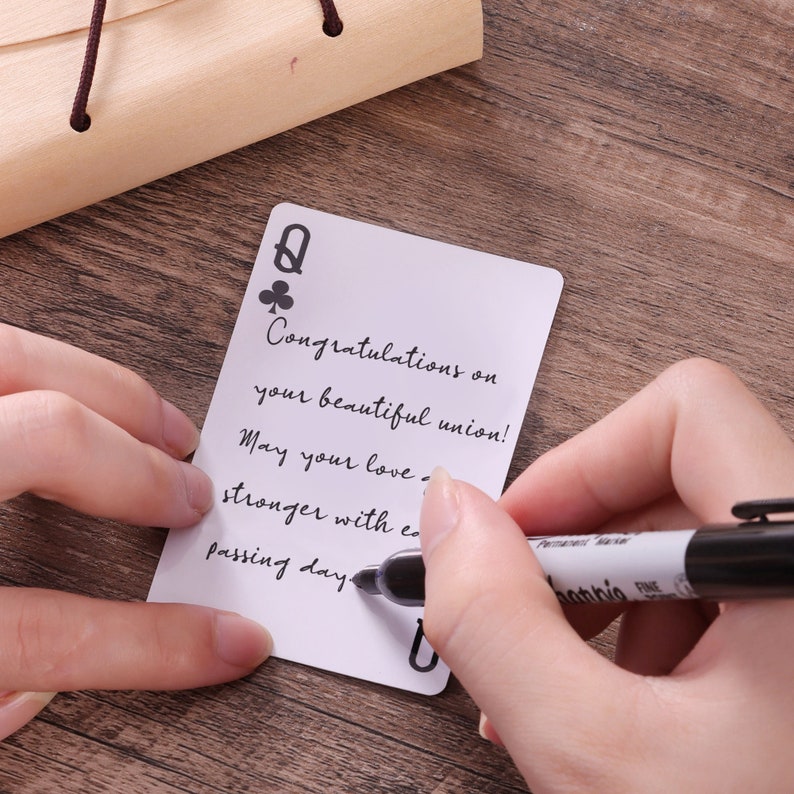 May include: A white rectangular card designed like a playing card with a club symbol and the letter Q in the corner. The card features the handwritten text: Congratulations on your beautiful union! May your love grow stronger with each passing day.