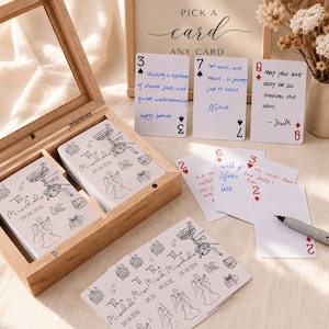 May include: Wooden box containing two decks of cards with a wedding theme. The cards feature illustrations of a bride and groom, disco balls, and the text "The Mischds 04.18.2026". Additional cards with handwritten messages and playing card suits are scattered around.