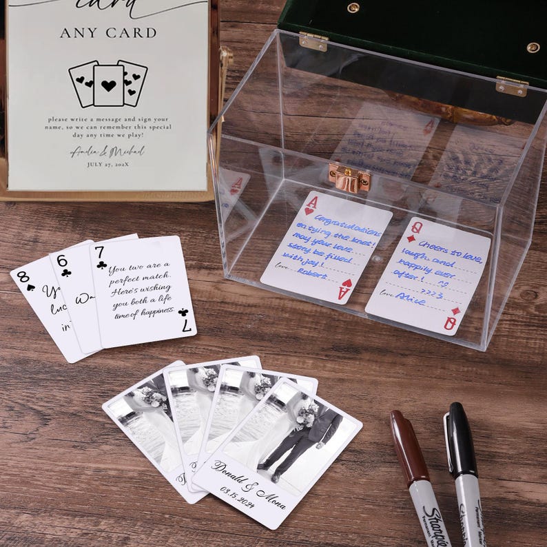 May include: A clear acrylic card box with a gold latch and dark green lid, accompanied by a sign reading ANY CARD, several white playing cards with handwritten messages, photo-printed cards featuring a couple, and two Sharpie markers.