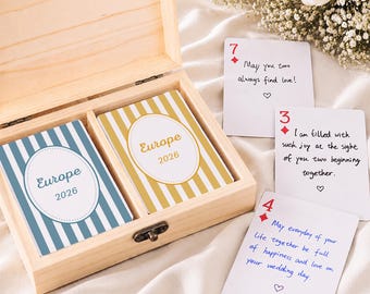 Può includere: Una scatola in legno chiaro contenente due mazzi di carte a righe blu e oro, con la scritta Europe 2026. Tre carte da gioco con semi di quadri rossi e messaggi scritti a mano sono poste accanto, ognuna con un simbolo di cuore.