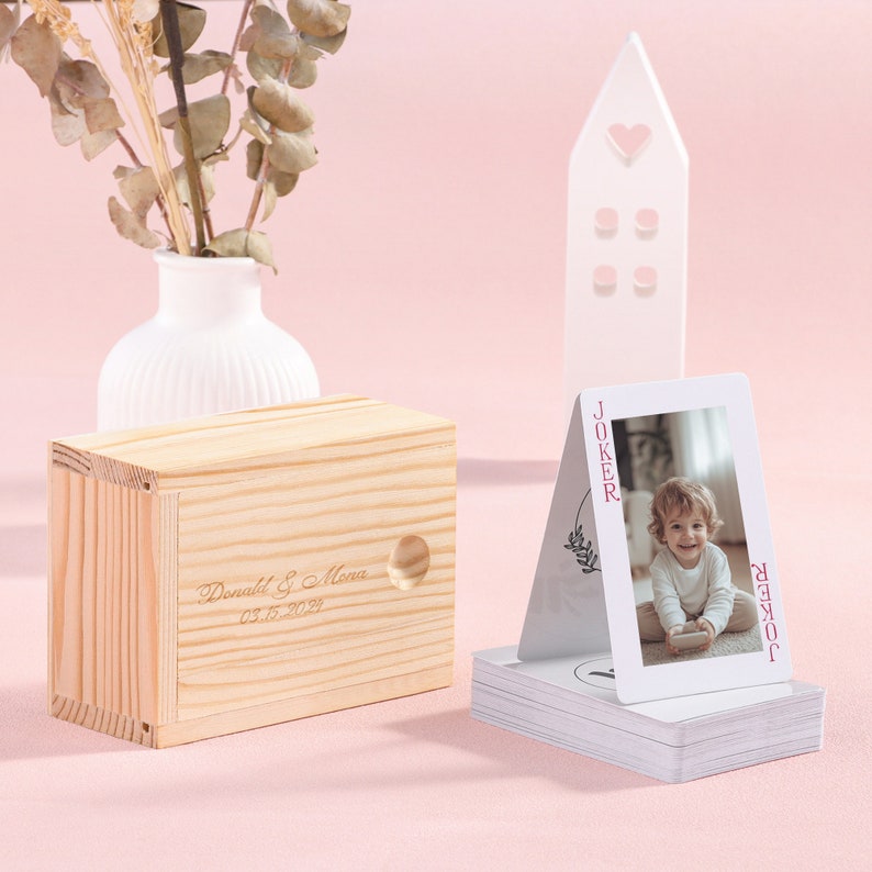 May include: A wooden box with a personalized inscription reading "Donald & Mena 03.15.2021" and a small hole in the top. A deck of playing cards with a custom joker card featuring a photo of a child is in the background.