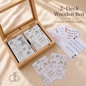 May include: A wooden box with a clear lid contains two decks of cards. The cards are decorated with wedding-themed illustrations and text, including names and dates. The box is made of light-coloured wood.