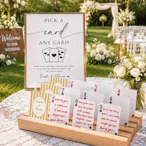 May include: A gold-framed sign with the text Pick a card any card and a wooden card holder displaying several playing cards with handwritten messages. The sign includes the names Derek and Olivia and a graphic of three playing cards.