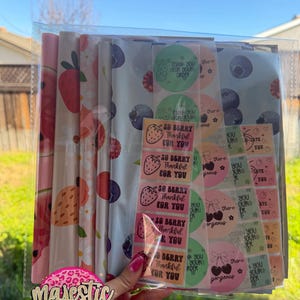May include: A clear plastic sleeve containing various packaging supplies. The supplies include patterned wrapping paper with fruit designs and stickers with phrases like "Thank You" and "So Berry Thankful For You". The "Majestic Mailers" logo is visible.