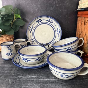 May include: A stoneware dinnerware set with a cream-colored base and blue leaf and line designs. The set includes a large plate, saucers, cups, and mugs. The mugs and cups have handles. The dinnerware is arranged on a dark wooden surface.