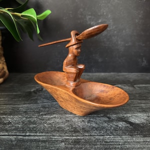 Puede incluir: Una escultura de madera tallada a mano que representa una figura sentada sosteniendo una cuchara, encima de un recipiente de doble cuenco. La escultura está hecha de madera marrón cálido, con un acabado liso. La figura lleva un sombrero.