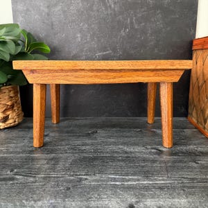 May include: A small, rectangular wooden riser with a natural light brown wood grain finish. The stand features four sturdy, slightly tapered legs supporting a flat, thick top surface, providing a stable platform for displaying various small items.