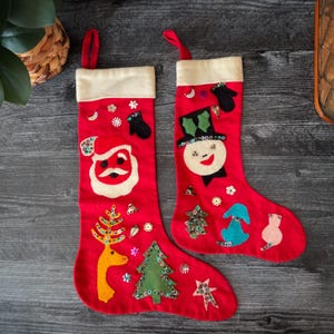 Könnte beinhalten: Zwei rote Weihnachtsstrümpfe mit weißen Bündchen und roten Schlaufen. Ein Strumpf zeigt ein Weihnachtsmann-Design, der andere einen Schneemann. Beide Strümpfe haben Filz- und Paillettenverzierungen, darunter Bäume, Sterne und Schneeflocken. Eine festliche Weihnachtsdekoration.