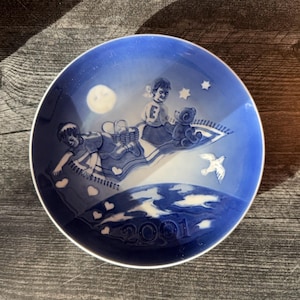 May include: A decorative blue and white plate featuring a whimsical scene of children on a flying carpet, a full moon, stars, and a dove. The plate has a white rim and the year "2001" is displayed at the bottom. A charming collectible.