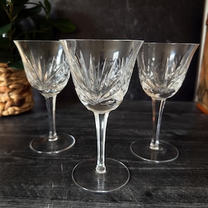 May include: Three clear crystal wine glasses with detailed starburst designs on the bowls. Each glass has a long stem and a circular base. The background is dark, with a hint of greenery and a woven basket.