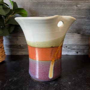 May include: A handmade ceramic vase with a unique design. The vase features a white top, a green and orange band, and a purple base. A gold-colored line runs down the side. The vase has a hole near the top and is suitable for displaying flowers.