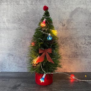 May include: A miniature Christmas tree with green needles, adorned with colorful lights, a red bow, and a red pom-pom at the top. The tree sits in a red pot and is decorated with holly and pinecones, creating a festive holiday display.