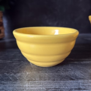以下が含まれることがあります： リブ加工が施された、小さくて丸い黄色のセラミックボウル。 ボウルは滑らかで光沢のある仕上がりで、わずかにフレアしたリムが付いています。 ボウルは暗い木製の表面に置かれています。