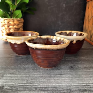 May include: Three brown ceramic bowls with a cream-colored rim. The bowls are arranged in a group, showcasing their rounded shape and textured exterior. The bowls are likely for serving or decorative purposes.