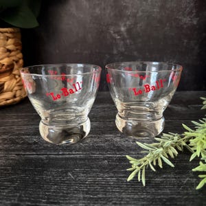 May include: Two clear, vintage-style lowball glasses with a rounded base. The glasses have a flared top and the words "Lo Ball" in red lettering. The glasses are on a dark wooden surface.