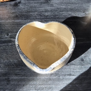 Może przedstawiać: Ceramiczna miska w kształcie serca z jasnobeżowym wnętrzem i niebiesko-szarą obwódką. Miska stoi na ciemnej drewnianej powierzchni, a światło słoneczne rzuca cienie. Kształt i kolor miski sugerują, że może być używana do serwowania lub dekoracji.