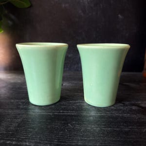 Puede incluir: Dos tazas de cerámica verde claro con un diseño acampanado. Las tazas tienen un acabado liso y brillante y una forma ligeramente curvada. Las tazas se exhiben sobre una superficie de madera oscura.