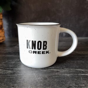 Peut inclure: Mug en céramique blanche avec des mouchetures noires et un bord noir. Le mug porte l'inscription "KNOB CREEK" en lettres noires grasses. La tasse a une anse incurvée et est posée sur un fond sombre.