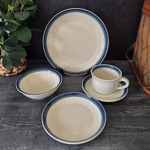 May include: A cream-colored ceramic dish set with blue and green trim. The set includes a large plate, a bowl, a smaller plate, a teacup, and a saucer. The dishes are arranged on a dark wooden surface.