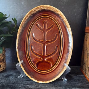 Könnte beinhalten: Ovale braune Keramikplatte mit einem erhabenen baumartigen Design in der Mitte. Die Platte hat einen dunkelbraunen Innenraum und einen helleren braunen, strukturierten Rand. Die Platte wird auf einem transparenten Kunststoffständer präsentiert.