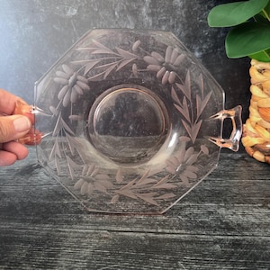 May include: A pink, octagonal glass serving dish with etched floral and leaf designs. The dish has a central circular indentation and two small handles. The design features daisies and foliage. The dish is displayed on a dark wooden surface.