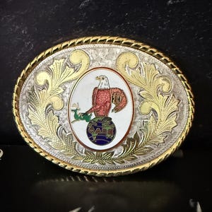 Puede incluir: Hebilla de cinturón ovalada plateada y dorada con un emblema de águila. La hebilla presenta un diseño floral ornamentado alrededor de los bordes y un óvalo central con un águila roja posada sobre un globo. Las letras "GABE" están en el globo.