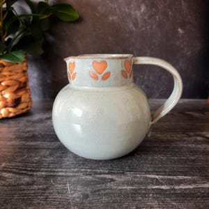 May include: A light blue ceramic pitcher with a rounded body and a curved handle. The rim is decorated with orange heart-shaped floral designs. The pitcher is likely used for serving beverages and adds a touch of vintage charm to any kitchen or dining setting.