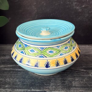 May include: A ceramic lidded jar featuring a light blue base with a ribbed lid. The body displays a horizontal band of green and blue diamond patterns above a yellow section decorated with dark blue vertical brushstroke accents and thin lines.