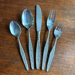 May include: A set of five silver-colored flatware pieces, including two forks, two spoons, and a knife. The handles feature a floral pattern. The utensils are arranged on a wooden surface.