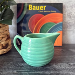 Puede incluir: Una cremera de cerámica turquesa con asa y boquilla. La cremera tiene un diseño acanalado alrededor del cuerpo. En el fondo hay un libro con el título "Bauer" y el subtítulo "Classic American Pottery".