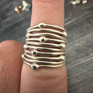Peut inclure: Une bague en argent au design unique. La bague présente plusieurs bandes d'argent, chacune avec une petite perle ronde. Les bandes sont empilées, créant un effet de superposition. La bague est portée au doigt.