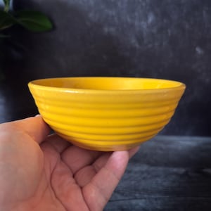 May include: A vibrant yellow ceramic bowl with a ribbed exterior. The bowl is held in a hand, highlighting its bright colour and design. The bowl's shape is round, and the interior is smooth.