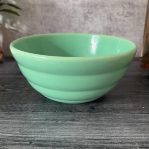 以下が含まれることがあります： ミントグリーンのセラミックボウル。丸みを帯びた形状で、外側に水平な溝があります。ボウルは空で、暗い木製の表面に置かれています。