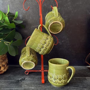 以下が含まれることがあります： 緑色のセラミックマグカップ4個セット。繰り返し模様の円のデザイン。赤い金属製のマグツリーに飾られています。マグカップには取っ手があり、日本製です。