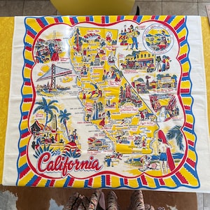 May include: A white, rectangular California souvenir tea towel with a colorful map design. The map features illustrations of landmarks and attractions, with the word "California" in a red script. The border is red, yellow, and blue.