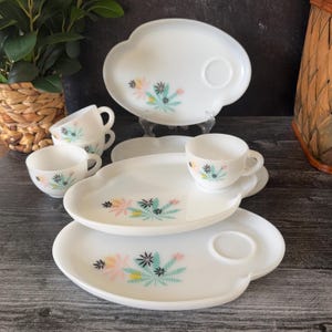 Puede incluir: Un juego de té vintage blanco con un diseño floral en rosa, turquesa y negro. El juego incluye platos, platillos y tazas de té. Los platos tienen forma de nube. Las tazas tienen asas. El juego está dispuesto sobre una superficie de madera oscura.