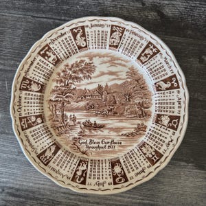 May include: A decorative plate with a brown and white landscape scene and a calendar for 1977. The plate features a scalloped edge and the text "God Bless Our House Throughout 1977". Zodiac signs and dates are printed around the edge.