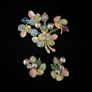 Könnte beinhalten: Ein Vintage-Schmuckset mit einer floralen Brosche und passenden Ohrringen. Die goldfarbenen Metallteile sind mit pastellfarbenem Emaille, Kunstperlen und funkelnden Strasssteinen verziert. Die Brosche und die Ohrringe haben ein zartes, feminines Design.