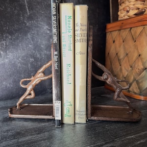 May include: Bronze-colored metal bookends with a figure design supporting a stack of books. The books include titles like "Indian Silversmithing" and "Navajo Shepherd and Weaver." The bookends and books are on a dark surface.