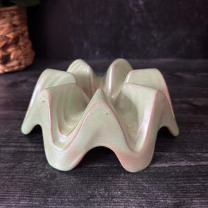 May include: A light green ceramic taco holder with a wavy design. The holder has a series of curved ridges, creating individual slots for tacos. The ceramic has a subtle, speckled texture and a hint of pinkish-brown along the edges.