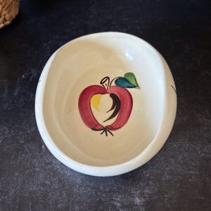 May include: Off-white ceramic bowl with a hand-painted apple design. The apple is red with a yellow center, a green leaf, and black details. The bowl has a rounded shape and a wide rim, suitable for serving food.