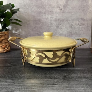Puede incluir: Una fuente de cerámica amarilla vintage con tapa, colocada en un soporte decorativo de metal dorado. El soporte presenta un diseño calado. La fuente tiene un acabado moteado y un pequeño pomo en la tapa.