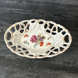 Puede incluir: Plato ovalado de cerámica blanquecina con diseño floral. El plato presenta un diseño calado en los bordes con ribete dorado. El centro del plato tiene un arreglo floral pintado en tonos morados, rojos y amarillos.