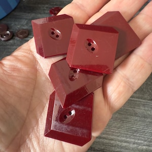 May include: A collection of burgundy square buttons with two holes each. The buttons have a faceted design and are held in a hand. Other smaller, round buttons are visible in the background.