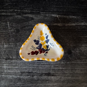Puede incluir: Un pequeño plato triangular de cerámica. Tiene una base blanca con lunares amarillos alrededor del borde. El plato está decorado con un motivo floral en amarillo, azul y rojo. Probablemente se utiliza para servir pequeñas porciones.