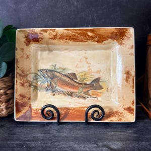 May include: A rectangular ceramic serving platter featuring a cream-colored center with a detailed illustration of a brown trout swimming among aquatic plants. The border has a distressed, textured brown finish. The signature Robert Gordon is visible.