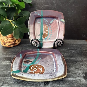 May include: Two square ceramic plates featuring a mottled purple and white glaze with abstract teal brushstrokes and brown circular motifs. The plates have a smooth, glossy finish and are displayed on a black metal stand for vertical viewing.