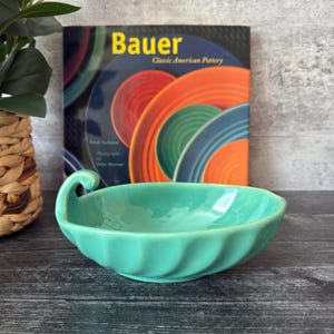 Puede incluir: Un cuenco de cerámica turquesa con borde festoneado y asa curvada. El cuenco está delante de un libro titulado "Bauer Classic American Pottery" con platos coloridos en la portada. El fondo es una superficie de madera oscura.